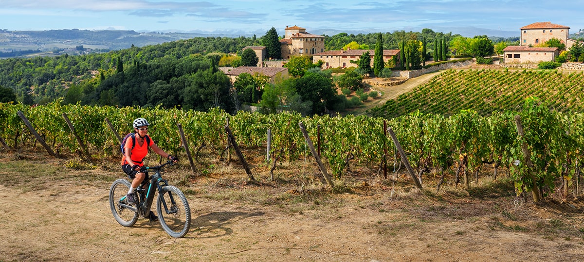 Cicloturismo enogastronomico donna in sella alla sua mountain bike elettrica tra vigne, ulivi e cipressi nella zona del Chianti in Toscana, Italia